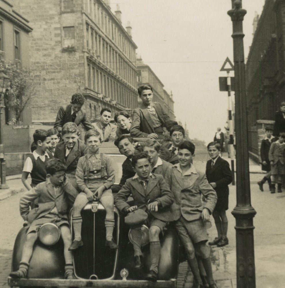 Black and white image of a group of boys sitting on a 1930s car on a Glasgow street