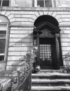 A black and white photographs shows a black door with glass squares in the centre. Surrounding the door is stonework, railings and foliage.