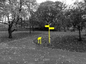 A black and white photograph of a paved path inside a park. It is the autumn and the trees are dropping their leaves. A wayfinding sign and map are blocked out in neon yellow.