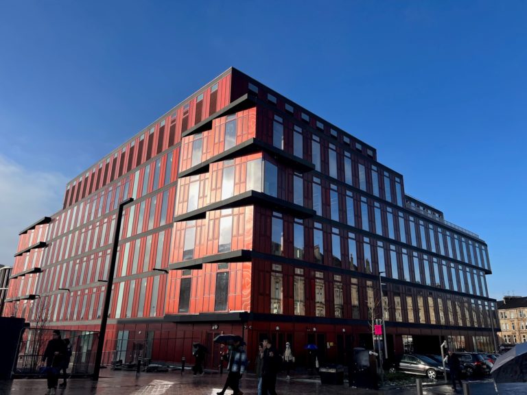 Large modern building in a rust red colour with lots of windows under a blue sky