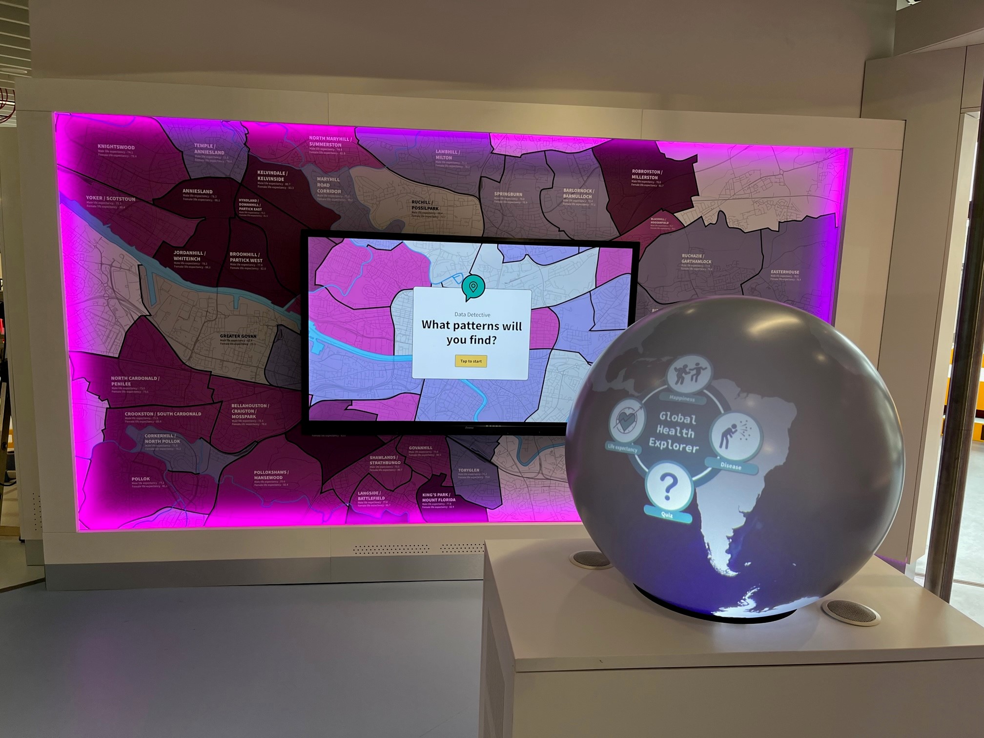 A lit up globe saying Global Health Explorer on a stand and behind a large map of Glasgow and a TV screen saying what pattern will you find? The installation is lit with a pink light around it.