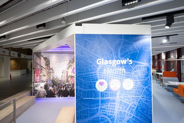 In the foreground there is a blue sign saying Glasgow's Health with infographic images of hands holding a heart, weighing scales and a lightblurb within a head outline and a map of Glasgow behind. In the background is a mosaic image of Buchanan Street in Glasgow and tables and shares in a an atrium area to the side