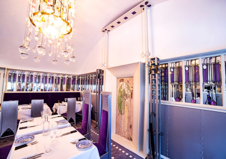 Image shows the interior of a tea saloon, designed by Charles Rennie Mackintosh. The top third of the walls is bright white, the centre third is clad with a band of decorative mirrors, the lower third is covered with silver fabric and glass beads. The room has a barrell vaulted ceiling, a large chandelier of clear, green and pink glass spheres, silver chairs upholstered with purple velvet, and a gesso panel displayed on the wall.
