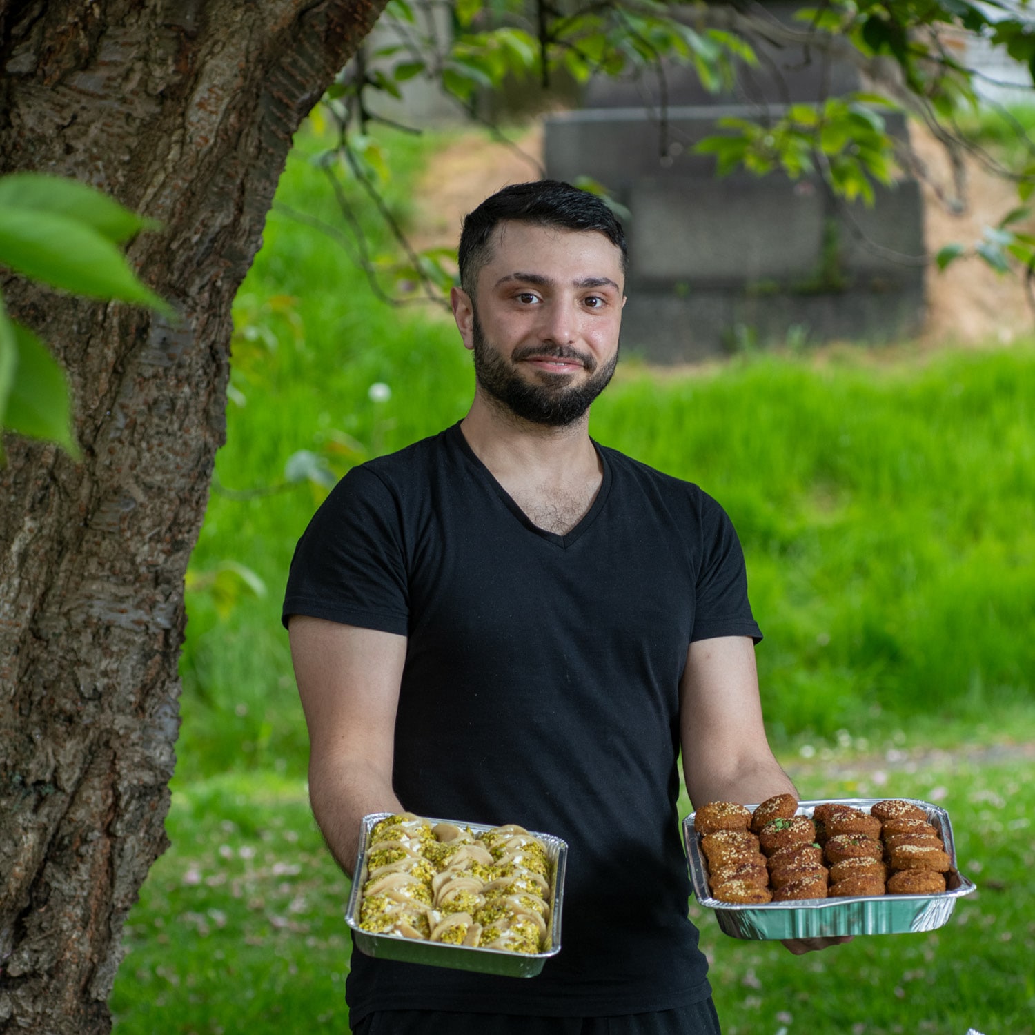 A man smiling in front of a tree holding out trays of food