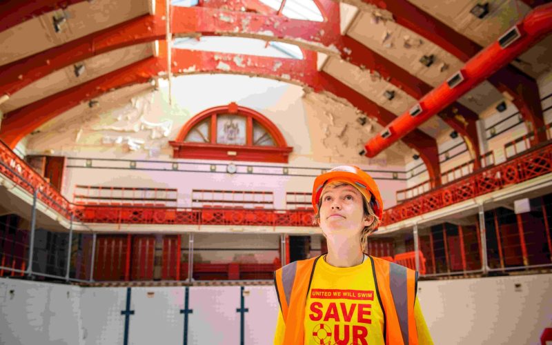 a woman wearing a hard hat, looking up at the ceiling of a swimming pool.